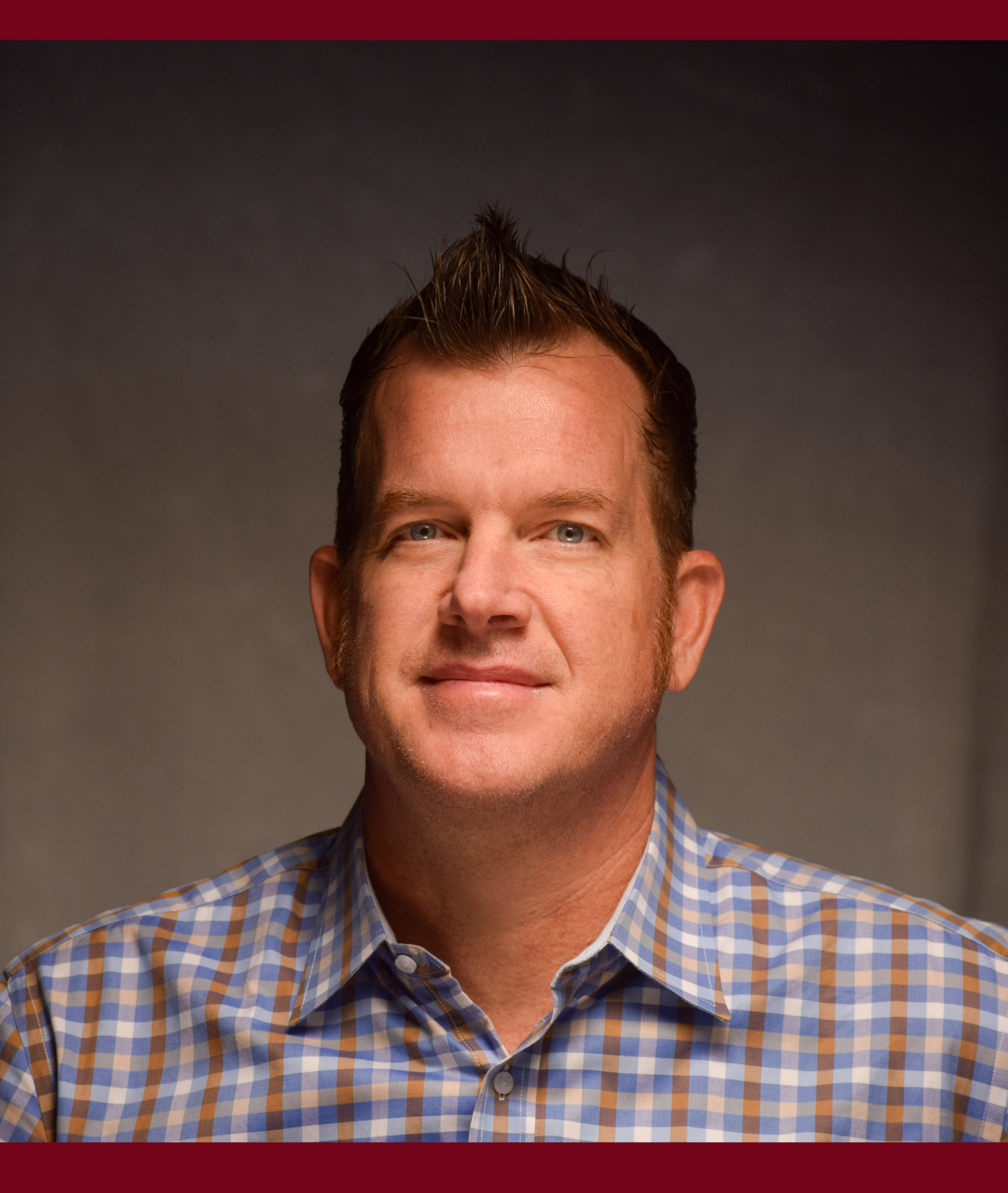 Smiling man in plaid shirt, photographed against gray backdrop with maroon top and bottom bars