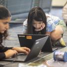 Two girls kneeling on carpet, focused on laptops and a small robot car on a mat.
